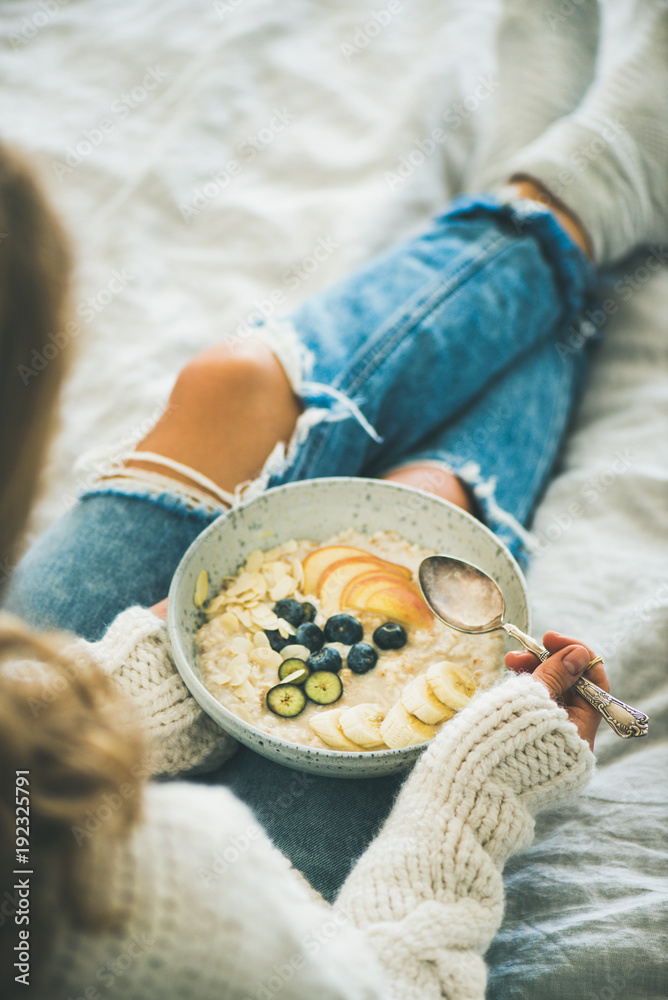 在床上吃健康的冬季早餐。穿着羊毛衫和破旧牛仔裤的女人在吃纯素食杏仁奶o