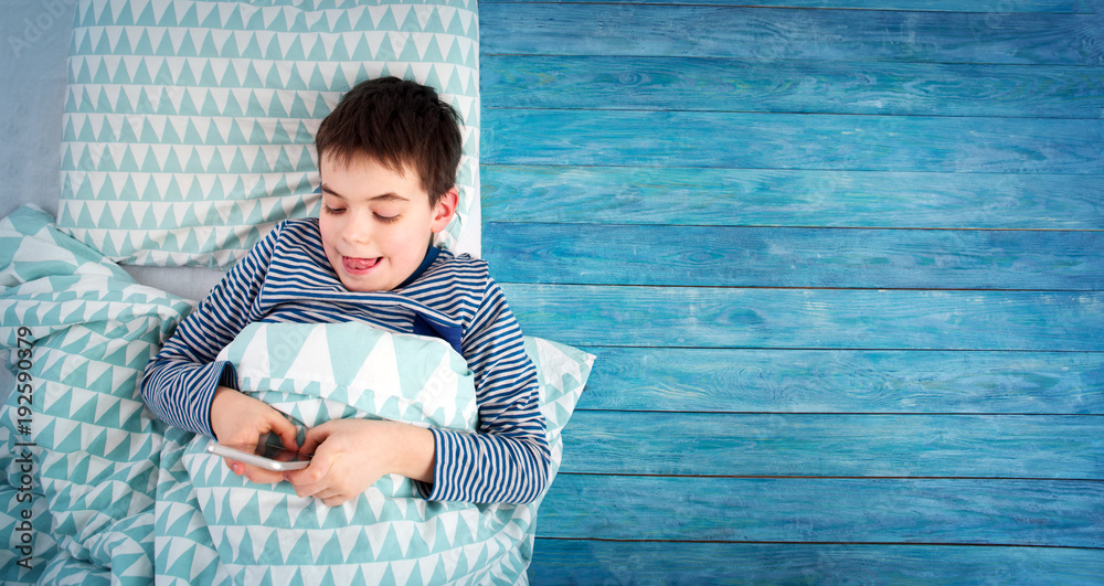 八岁的孩子睡在床上的枕头上。男孩躺在蓝色木制背景下，带智能手机。