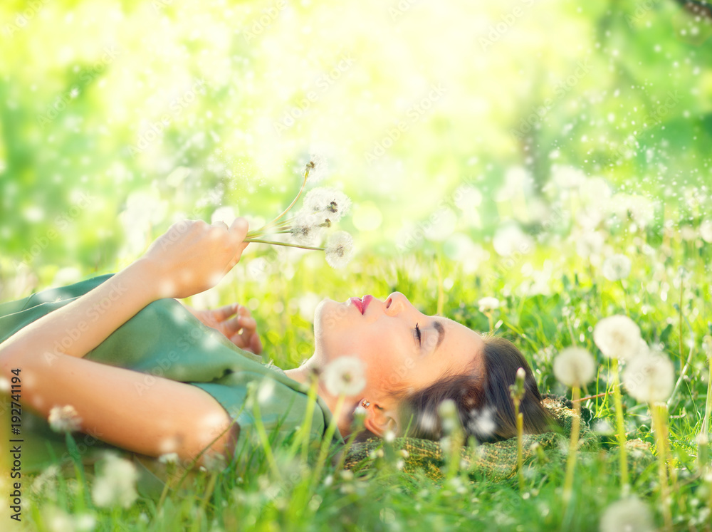 美丽的年轻女子躺在绿草上吹蒲公英。无过敏概念