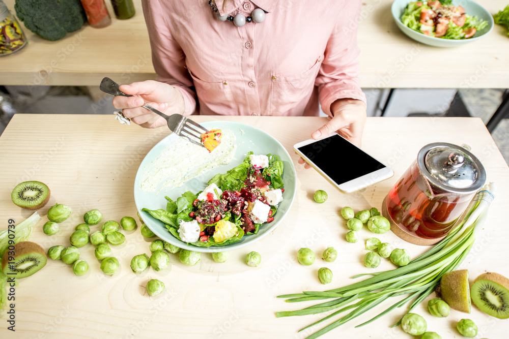 女人在餐桌上吃沙拉，用智能手机装饰绿色食品配料。俯视图
