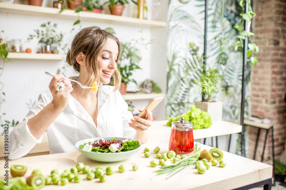 年轻女性坐在开着绿色花朵的美丽室内，拿着智能手机吃着健康的食物
