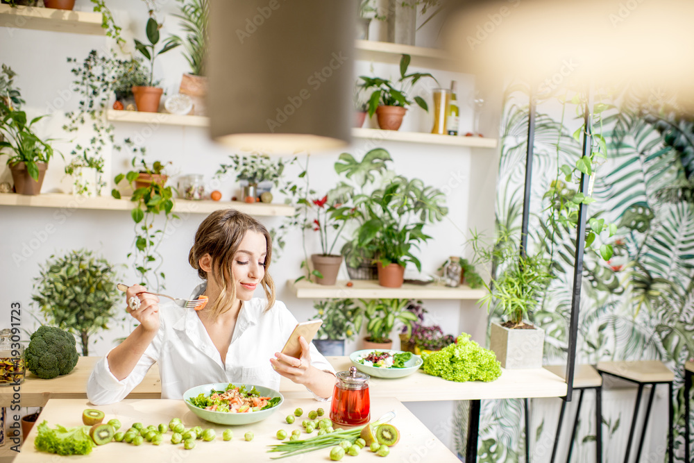 年轻女子坐在美丽的室内吃健康食品，背后开着绿色的花朵