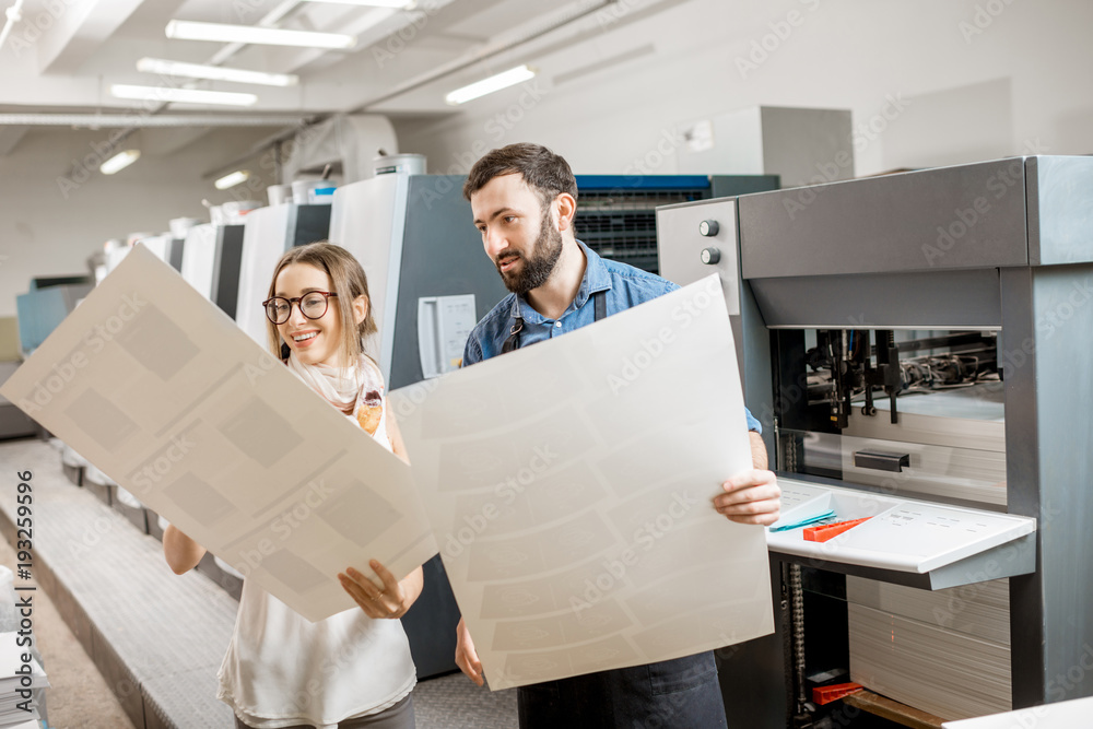 年轻的女性设计师和印刷操作员在印刷制造厂与纸印刷一起工作