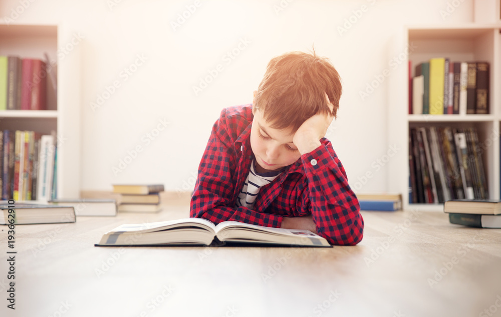 八岁的孩子躺在家里的地板上看书。严肃的男孩在白色房间里看书