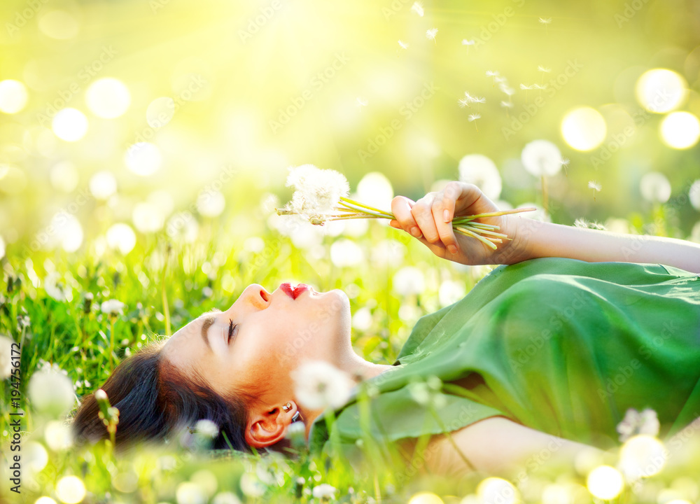 美丽的年轻女子躺在绿草中的田野上，吹着蒲公英花。无过敏