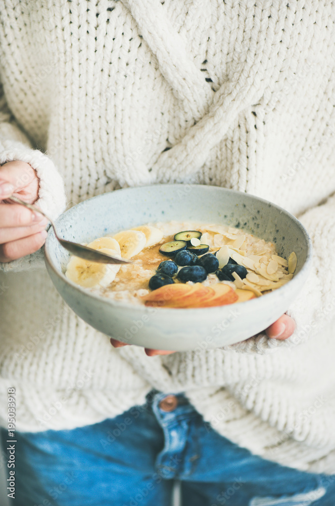 Healthy winter breakfast in bed. Woman in woolen sweater and jeans eating vegan almond milk oatmeal 