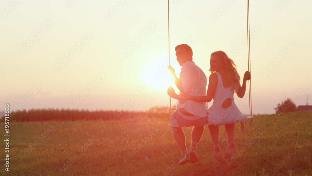 LENS FLARE: Lovely young smiling couple swinging in the summer sunset in nature.