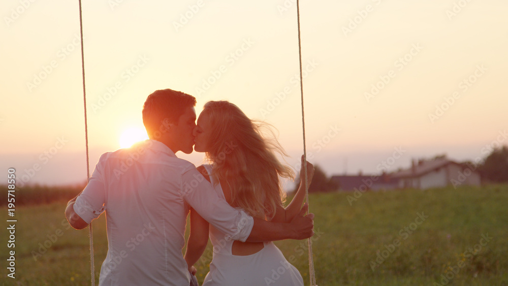 LENS FLARE CLOSE UP: Beautiful couple sharing a kiss while sitting on rope swing