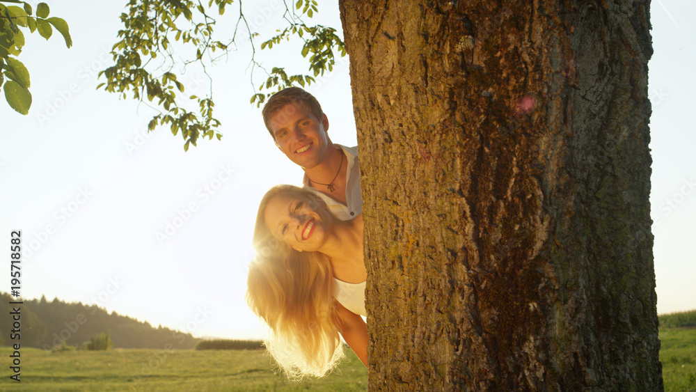 特写美丽的年轻夫妇从树后偷看，对着镜头微笑
