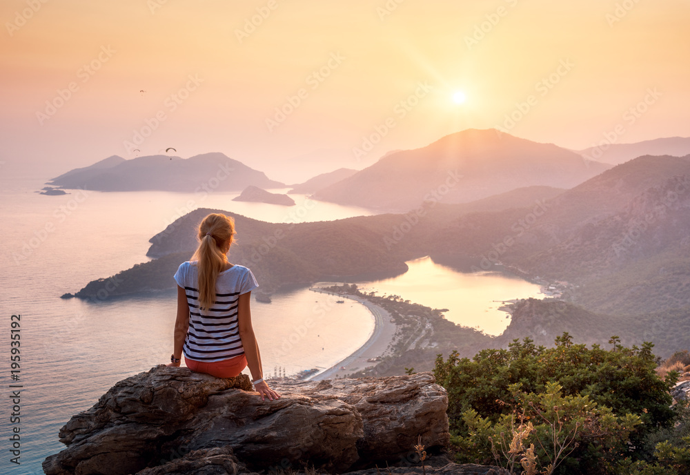 年轻女子坐在岩石顶部，在五彩缤纷的日落中眺望海岸和山脉
