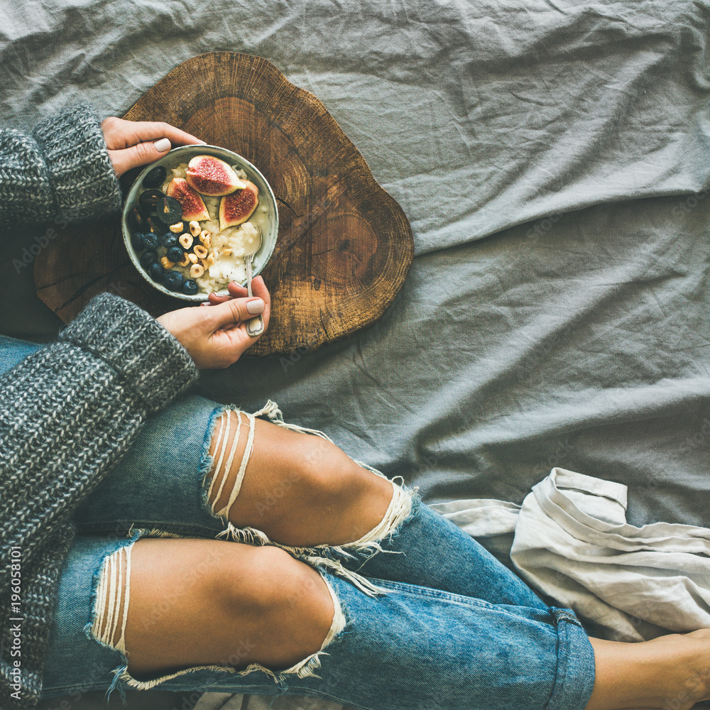 健康的冬季早餐在床上。穿着灰色毛衣和牛仔裤的女人在吃米饭椰子粥