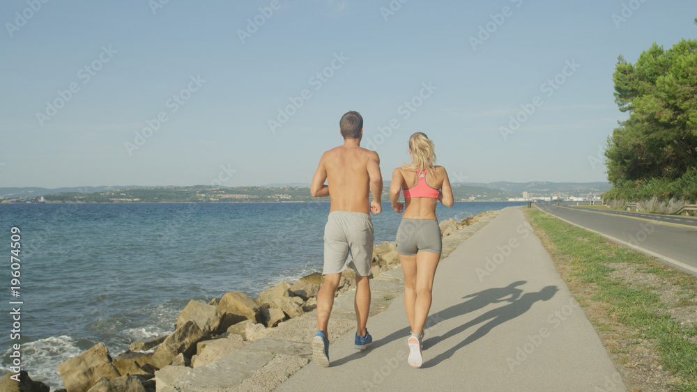 在阳光明媚的日子里，一对不知名的运动型男女沿着空旷的海洋道路奔跑。