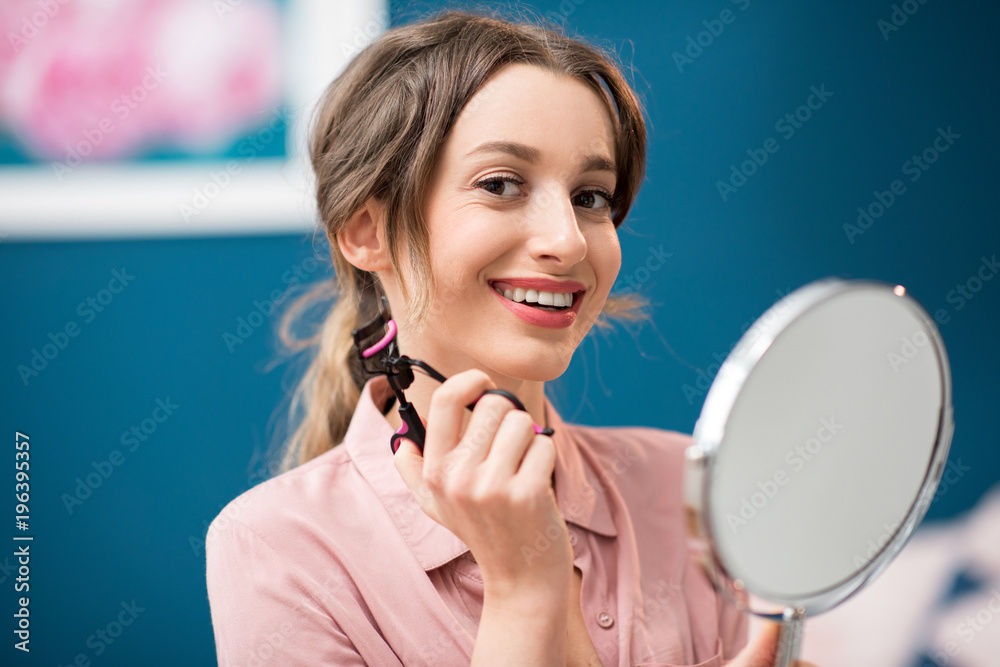 室内蓝色墙壁背景上坐着镜子，用睫毛夹的年轻女子