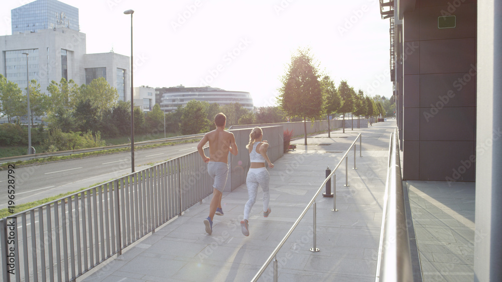 金发女子和赤膊男子沿着斜坡向绿色大道慢跑。