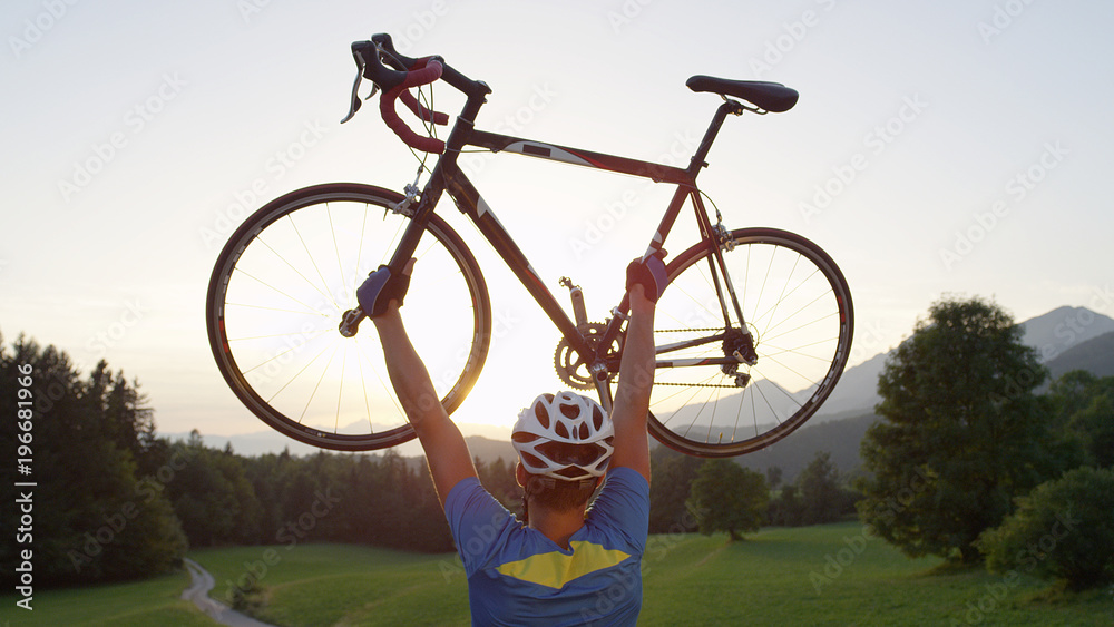 CLOSE UP: Ecstatic road biker lifting his bike over head while looking at sunset