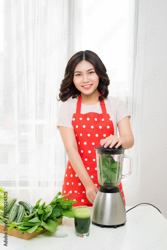 年轻微笑的女人在家里厨房的搅拌机里用新鲜的蔬菜做奶昔。健康的蔬菜