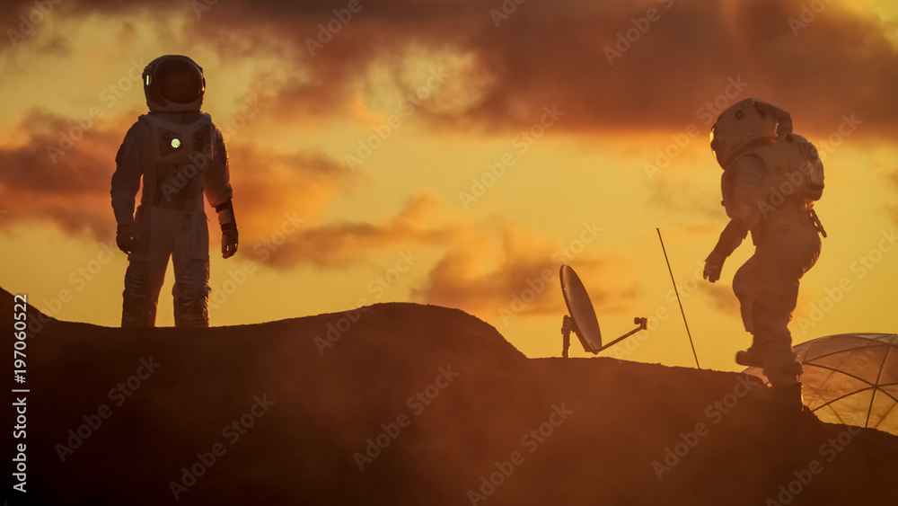 Two Silhouettes of the Astronauts Explore Red Rocky Alien Planet. In the Background Sunset with Base
