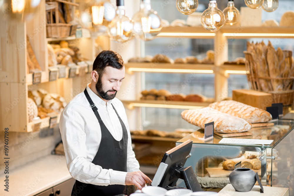 Handsome bread seller working with cash register in the small and cozy store with bakery products