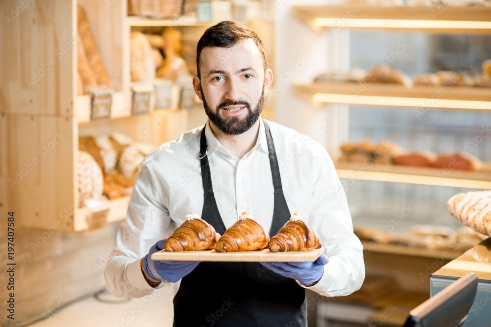 Portrait of a handsome seller in uniform standing with delicious croissants in the store with bakery