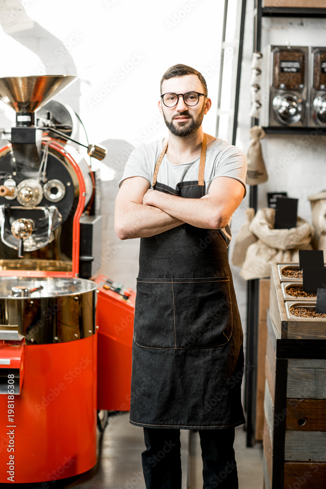 一位穿着制服的英俊咖啡师站在咖啡馆里的肖像，咖啡馆的ba上有烘焙机