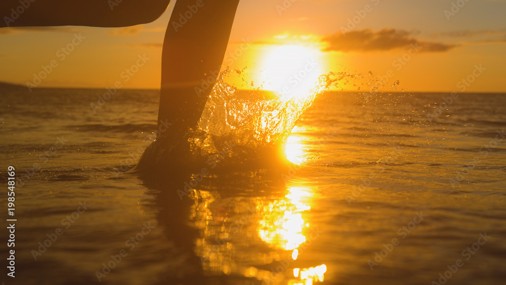 特写：一位无忧无虑的女士在金色日落时赤脚在浅水中奔跑。