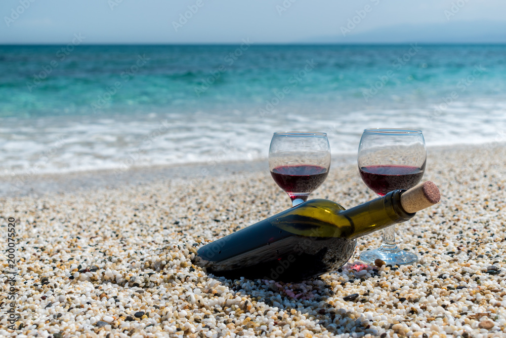 夏日阳光明媚的海滩上，一杯杯红酒和一瓶葡萄酒。背景是大海。