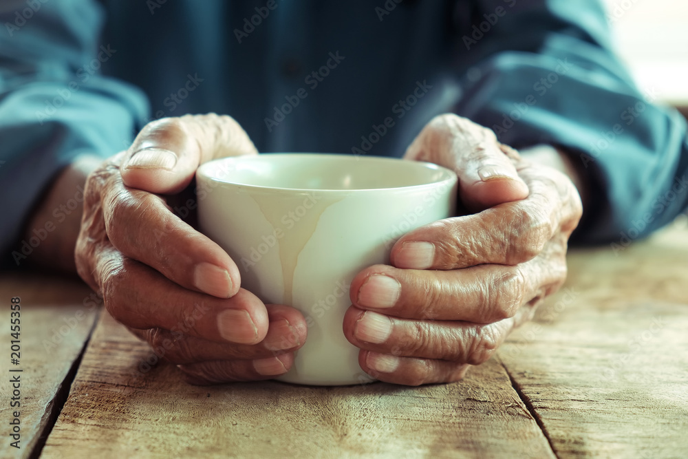 老人的手拿着一杯咖啡放在木桌上。语调