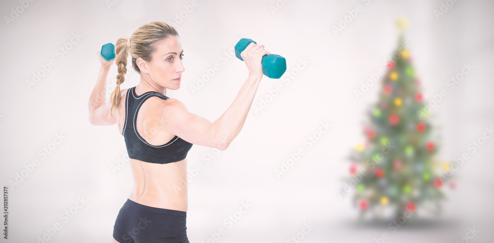 女健美运动员双手举着两个哑铃，靠着房间里模糊的圣诞树