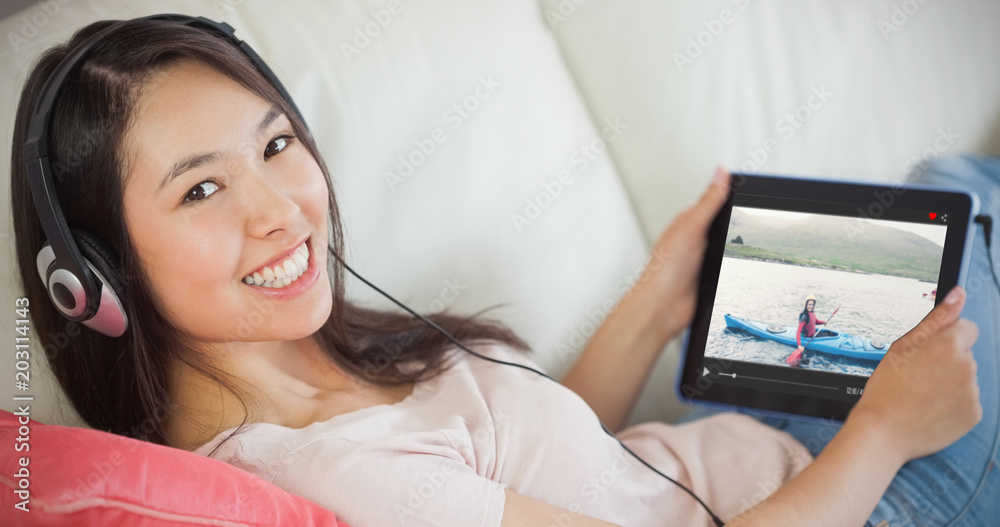 Girl using her tablet pc on the sofa and listening to music smiling at camera against smiling woman 