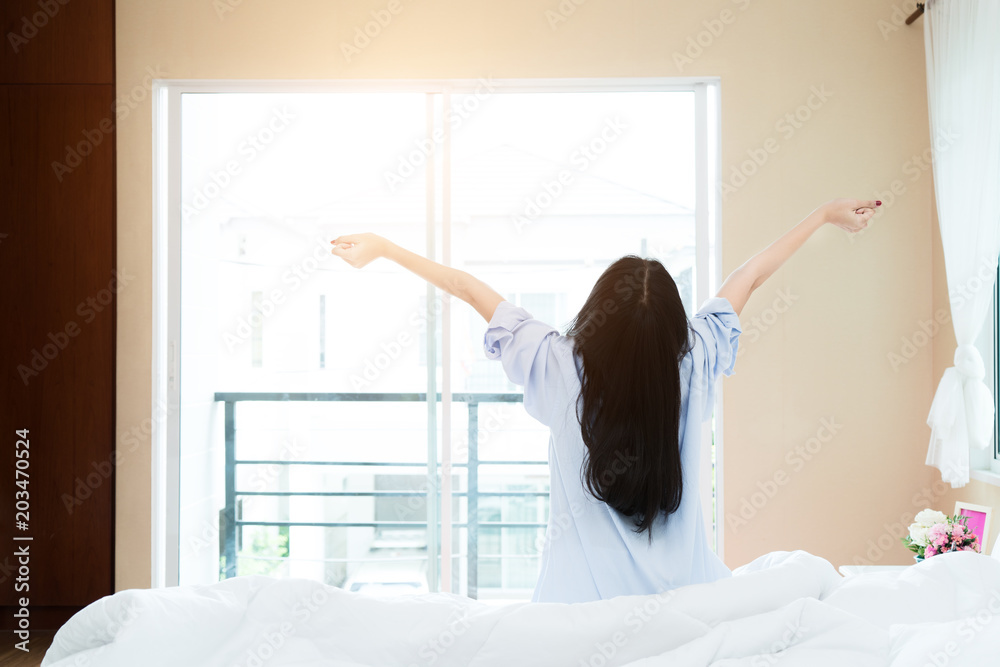 Back view of Asian woman stretching in bed after wake up, rising hands to window in the morning with
