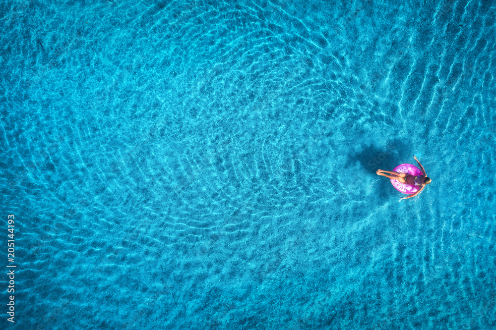 日出时，一名年轻女子在透明的蓝色海洋中的粉色游泳圈上游泳的鸟瞰图。苏