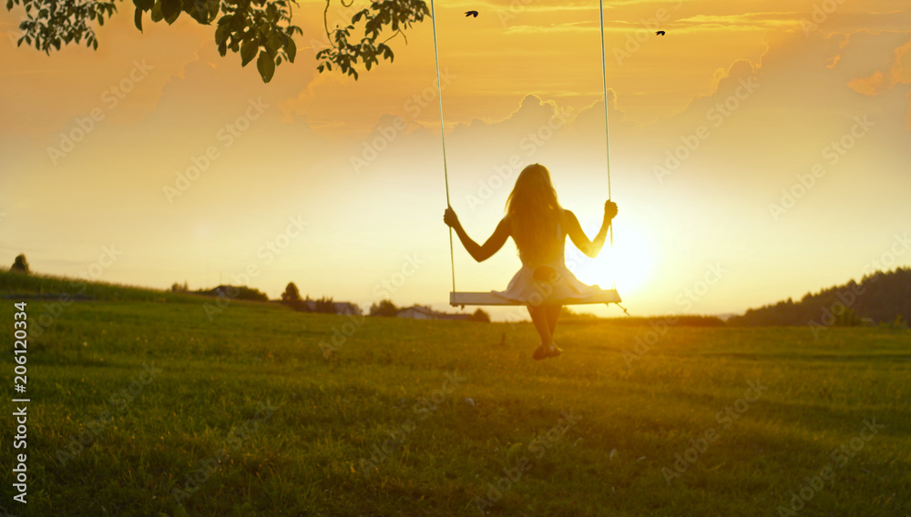 SILHOUETTE：金色夏日日落时，一个无法辨认的女孩在树上荡秋千
