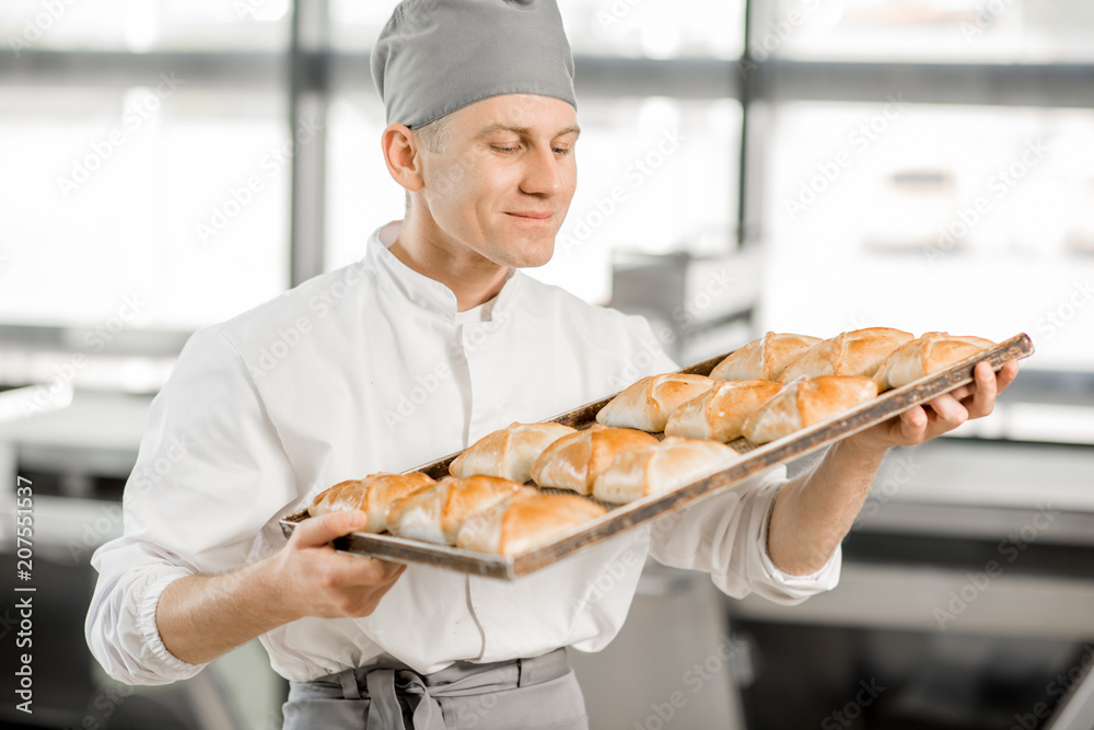 一个英俊的面包师的画像，托盘里装满了新鲜出炉的面包，站在面包厂