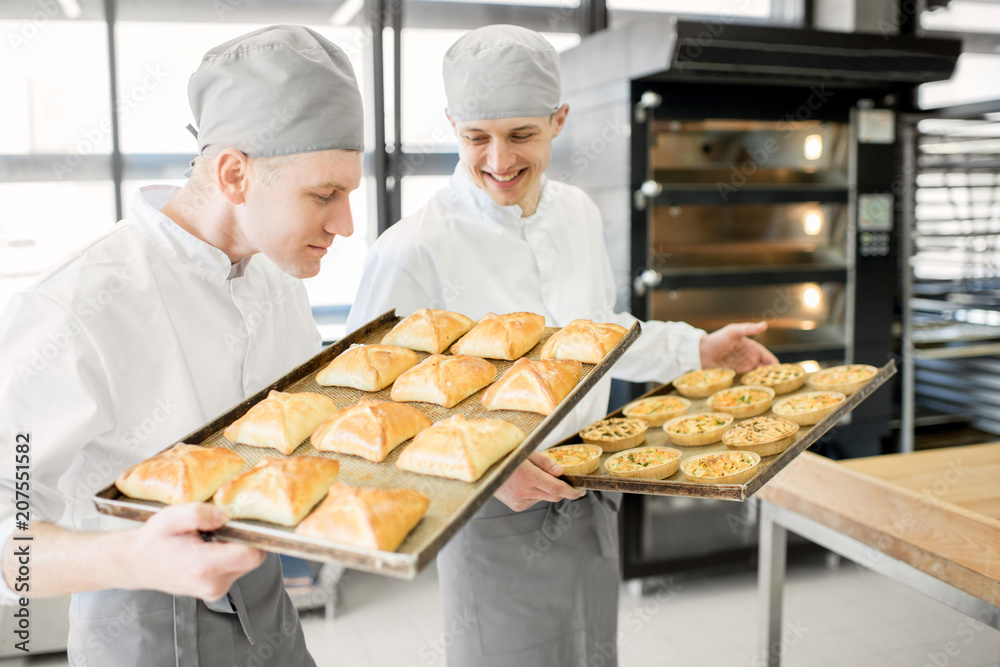 两位英俊的面包师站在生产现场，托盘里装满了新鲜出炉的面包