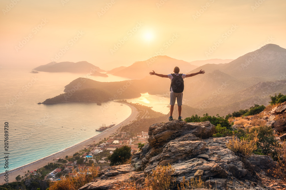 快乐的运动型男人，背着背包，举起双臂，站在岩石上看着大海
