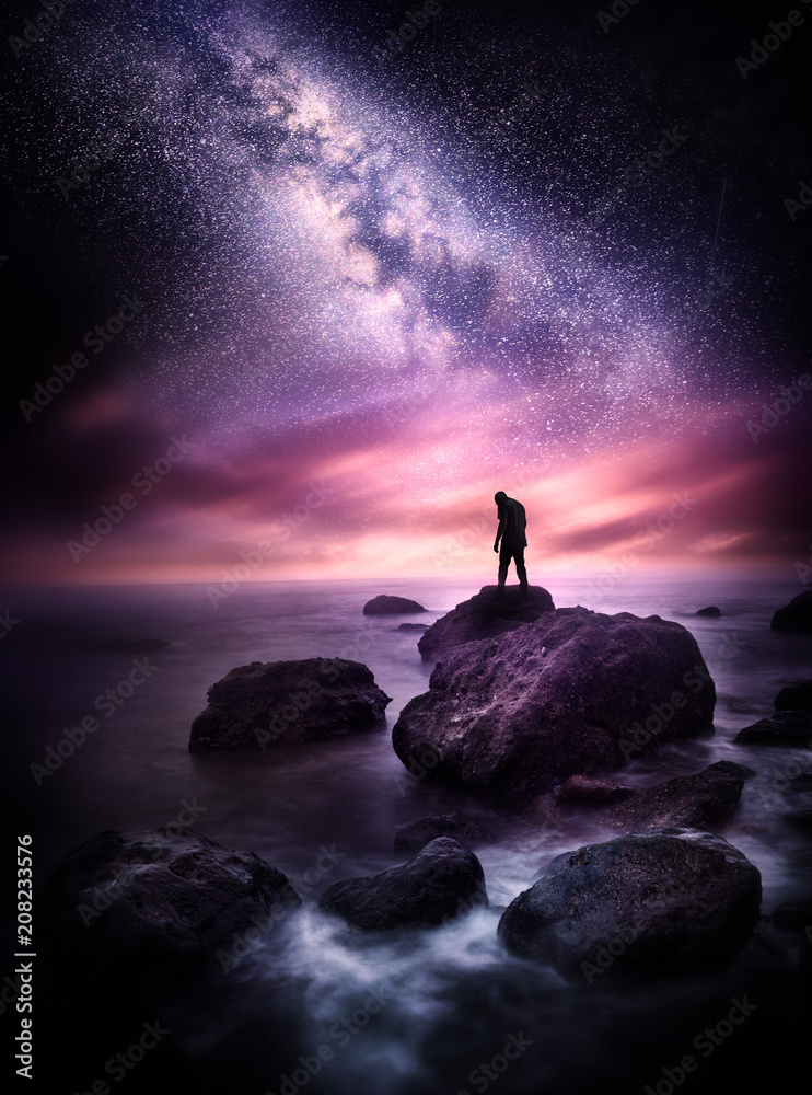 银河系的夜间海洋景观。一个人站在岩石海岸线上，上面有星星
