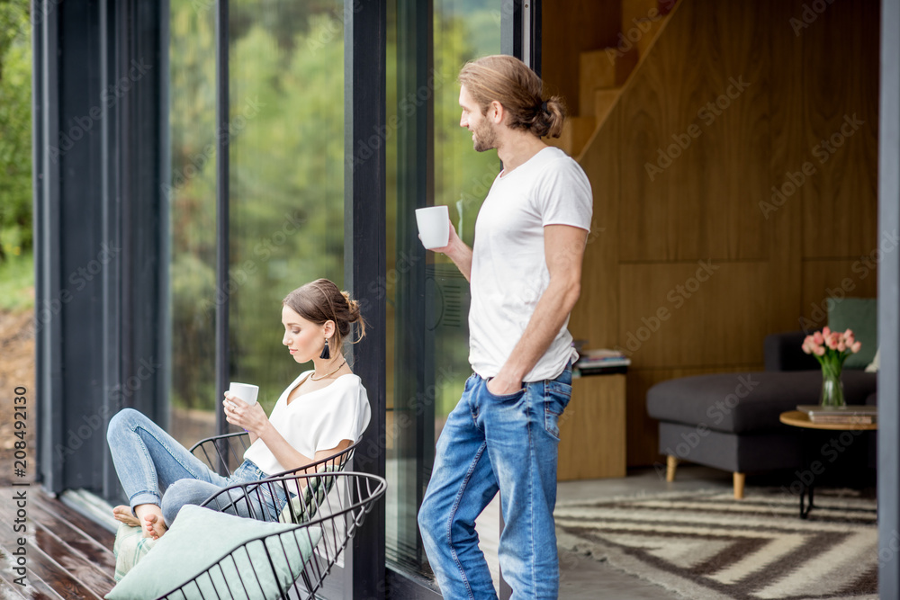年轻夫妇端着杯子坐在现代房子的露台上，欣赏户外美景