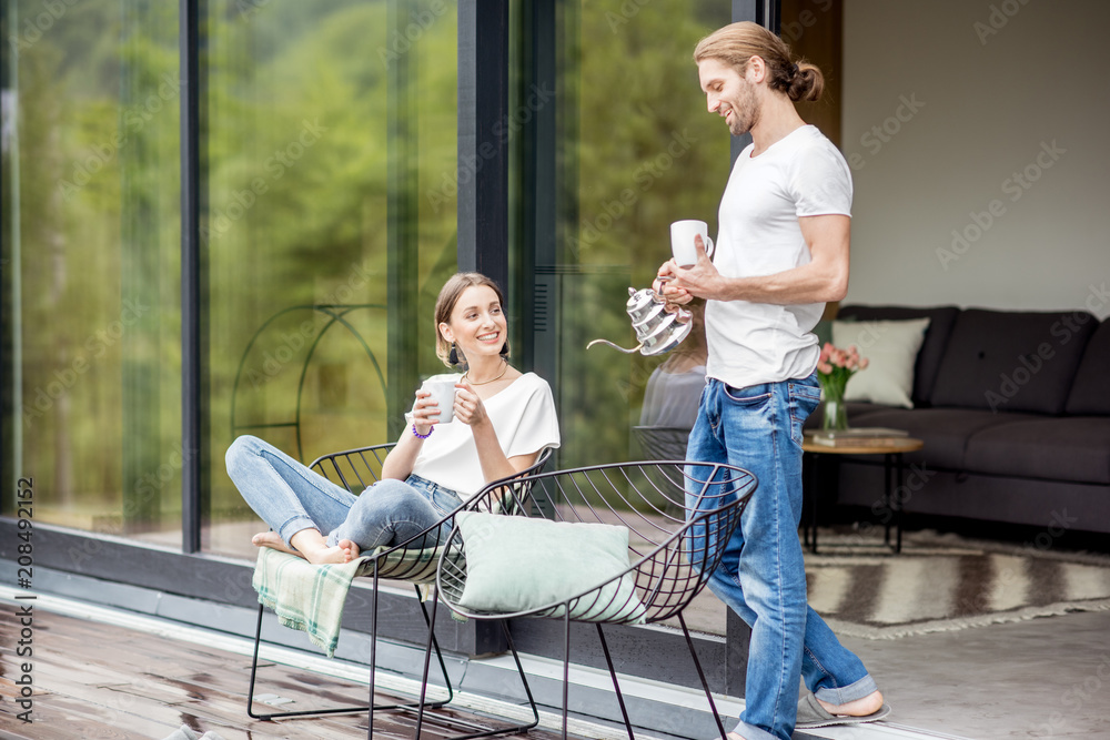 年轻夫妇端着杯子坐在现代房子的露台上，欣赏户外美景