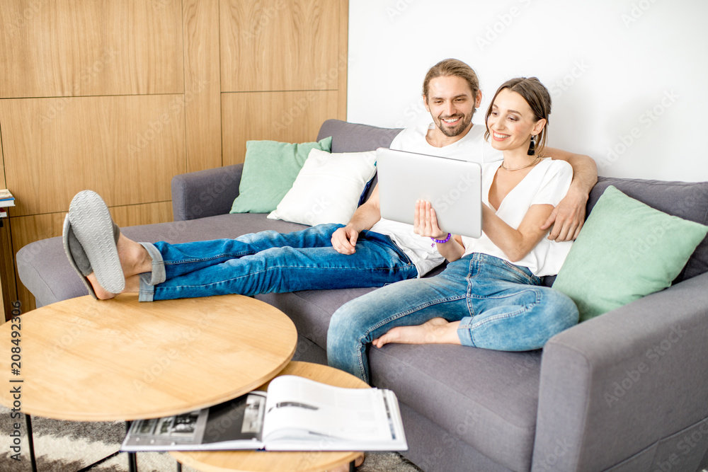 一对年轻夫妇穿着白衬衫和牛仔裤，使用数字平板电脑坐在沙发上