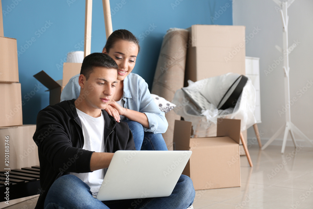一对年轻夫妇带着笔记本电脑坐在室内盒子附近的地板上。搬进新房子