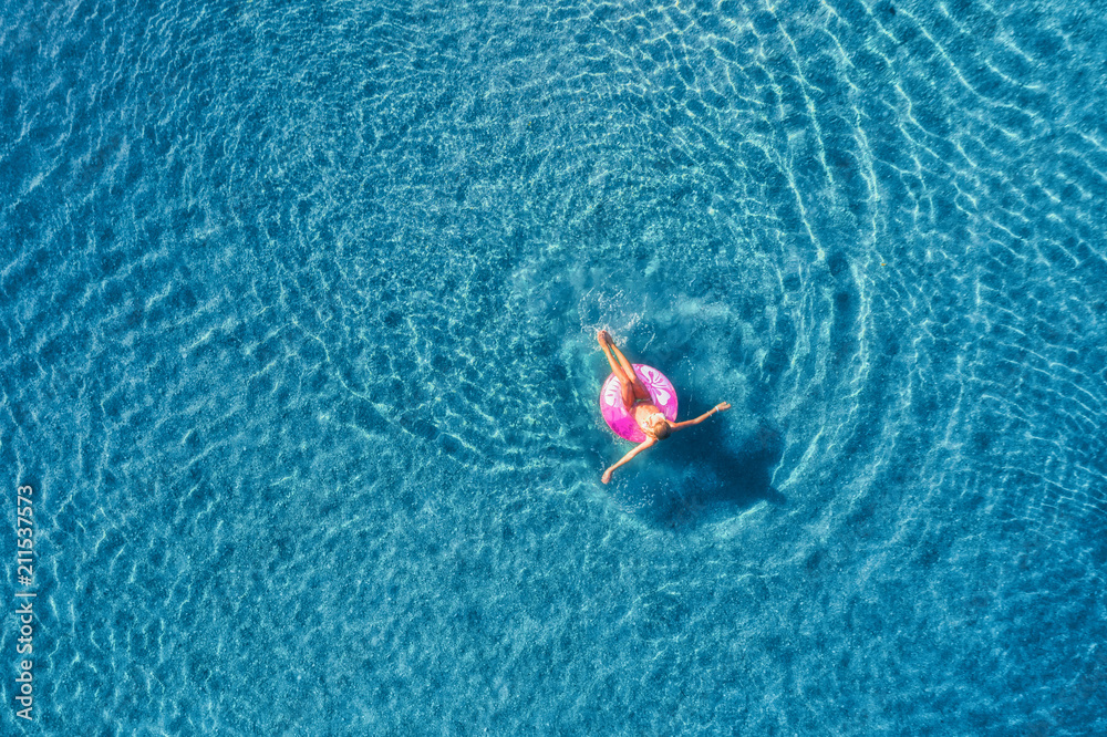晴朗的日子里，一名年轻女子在透明的蓝色海洋中的粉色游泳圈上游泳的鸟瞰图。