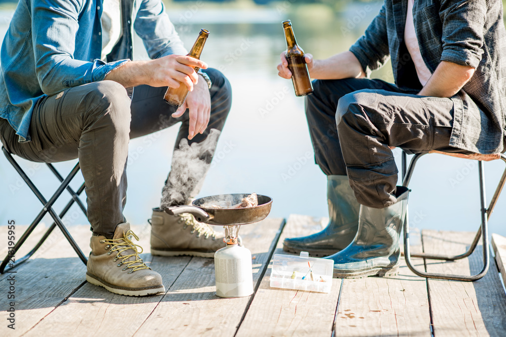 两名渔民在湖附近的木码头上野餐时坐着用啤酒煎鱼