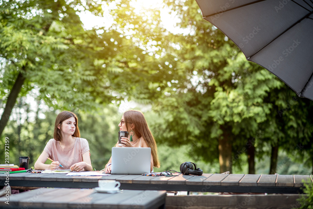 年轻的女朋友们坐在一起，拿着笔记本电脑和咖啡在户外公园咖啡馆学习