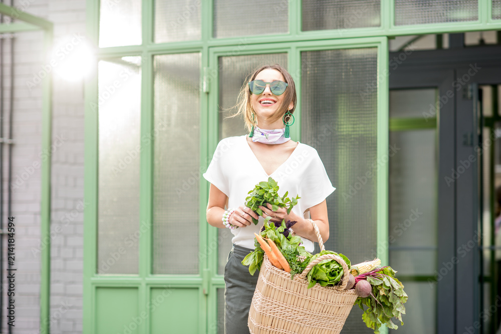 一位年轻女子的画像，她拿着装满新鲜生蔬菜的袋子，站在户外的绿色墙壁附近