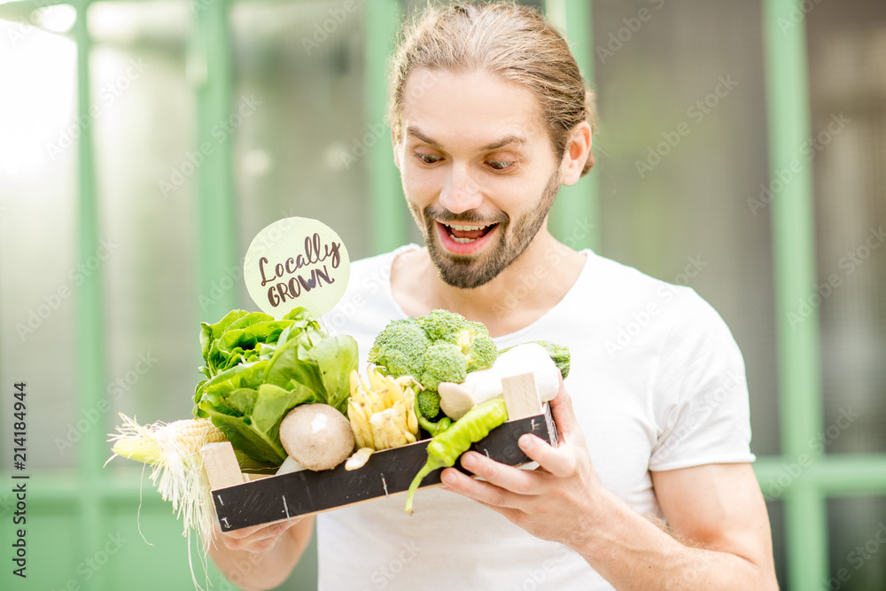 一个快乐的素食主义者的画像，他在户外的绿色ba上拿着装满新鲜生蔬菜的盒子