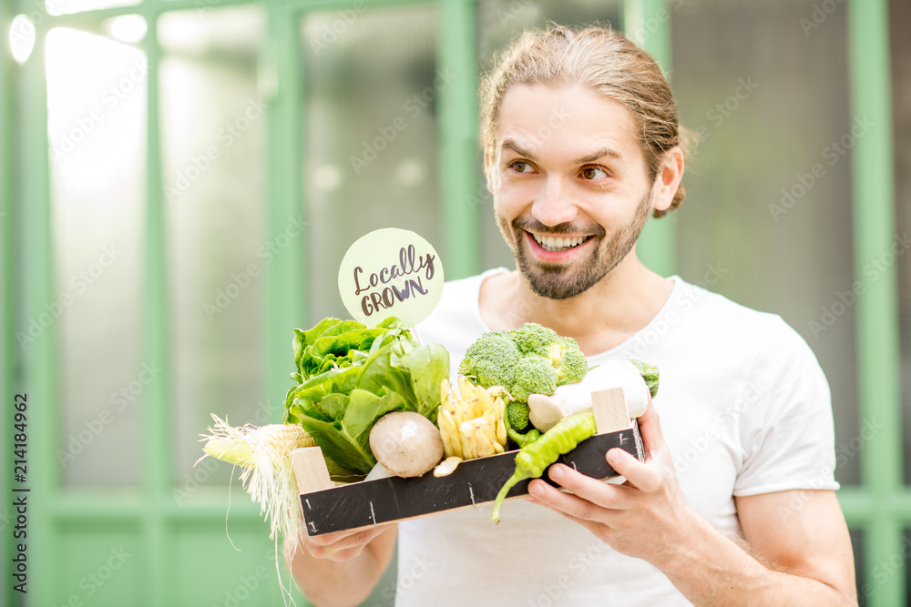 一个快乐的素食主义者的肖像，他在户外的绿色草坪上拿着装满新鲜生蔬菜的盒子