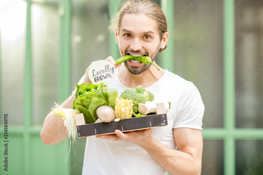 一个快乐的素食者的肖像，他拿着装满新鲜生蔬菜的盒子，在户外咬着辣椒