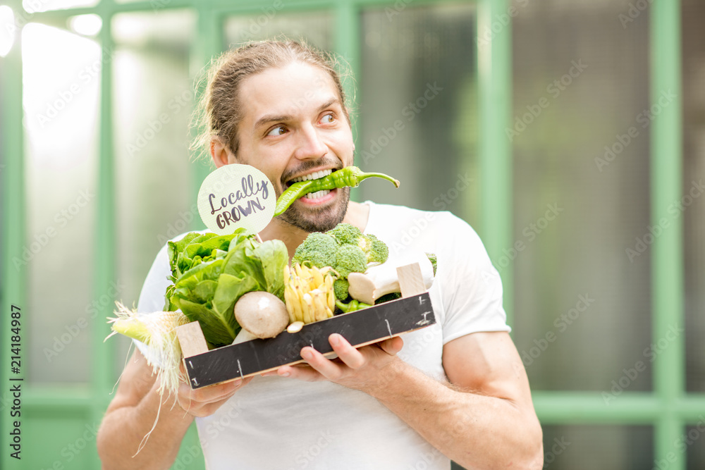 一个快乐的素食者的肖像，他拿着装满新鲜生蔬菜的盒子，在户外咬着辣椒