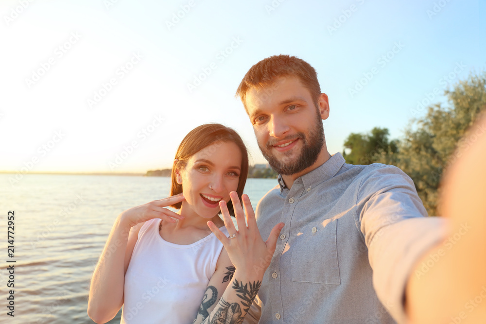 阳光明媚的日子里，一对幸福的情侣在河边拿着订婚戒指自拍