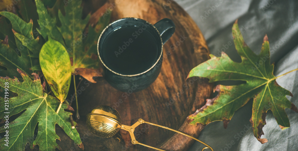 秋天的早茶躺在床上。一杯带筛子的红茶和五颜六色的落叶放在乡村的木制托盘上